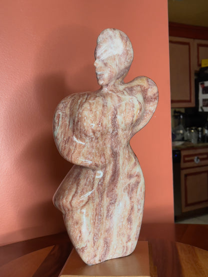 A hand-carved Utah alabaster sculpture depicting Venustus (pronounced veh-NUSS-tuss), the Goddess of Love, showcasing an elegant and refined female form. It represents the qualities of beauty, confidence, and self-assurance, embodying a woman who confidently navigates her own journey through life. The sculpture reflects themes of independence and personal strength, celebrating resilience and empowerment.

Venustus is an original design created by Shahla Rahimi Reynolds.

The piece is 13" H x 8" W x 3.5" D.