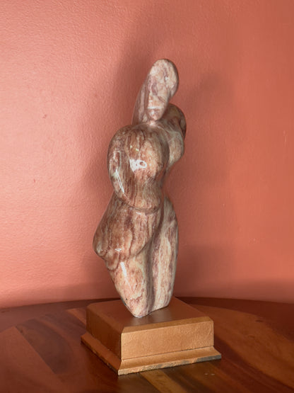 A hand-carved Utah alabaster sculpture depicting Venustus (pronounced veh-NUSS-tuss), the Goddess of Love, showcasing an elegant and refined female form. It represents the qualities of beauty, confidence, and self-assurance, embodying a woman who confidently navigates her own journey through life. The sculpture reflects themes of independence and personal strength, celebrating resilience and empowerment.

Venustus is an original design created by Shahla Rahimi Reynolds.

The piece is 13" H x 8" W x 3.5" D.