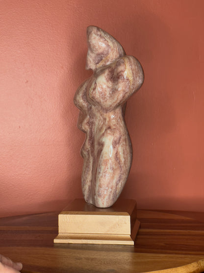 A hand-carved Utah alabaster sculpture depicting Venustus (pronounced veh-NUSS-tuss), the Goddess of Love, showcasing an elegant and refined female form. It represents the qualities of beauty, confidence, and self-assurance, embodying a woman who confidently navigates her own journey through life. The sculpture reflects themes of independence and personal strength, celebrating resilience and empowerment.

Venustus is an original design created by Shahla Rahimi Reynolds.

The piece is 13" H x 8" W x 3.5" D.